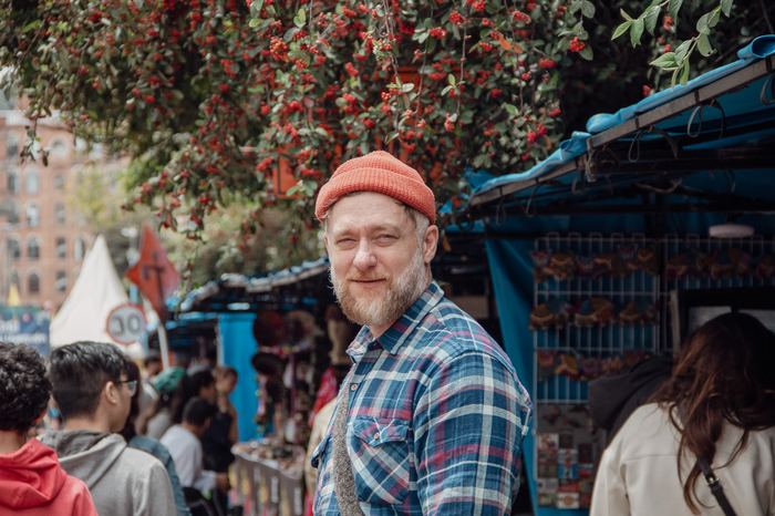 A person in a red beanie and plaid shirt, standing at an outdoor market under a berry tree.